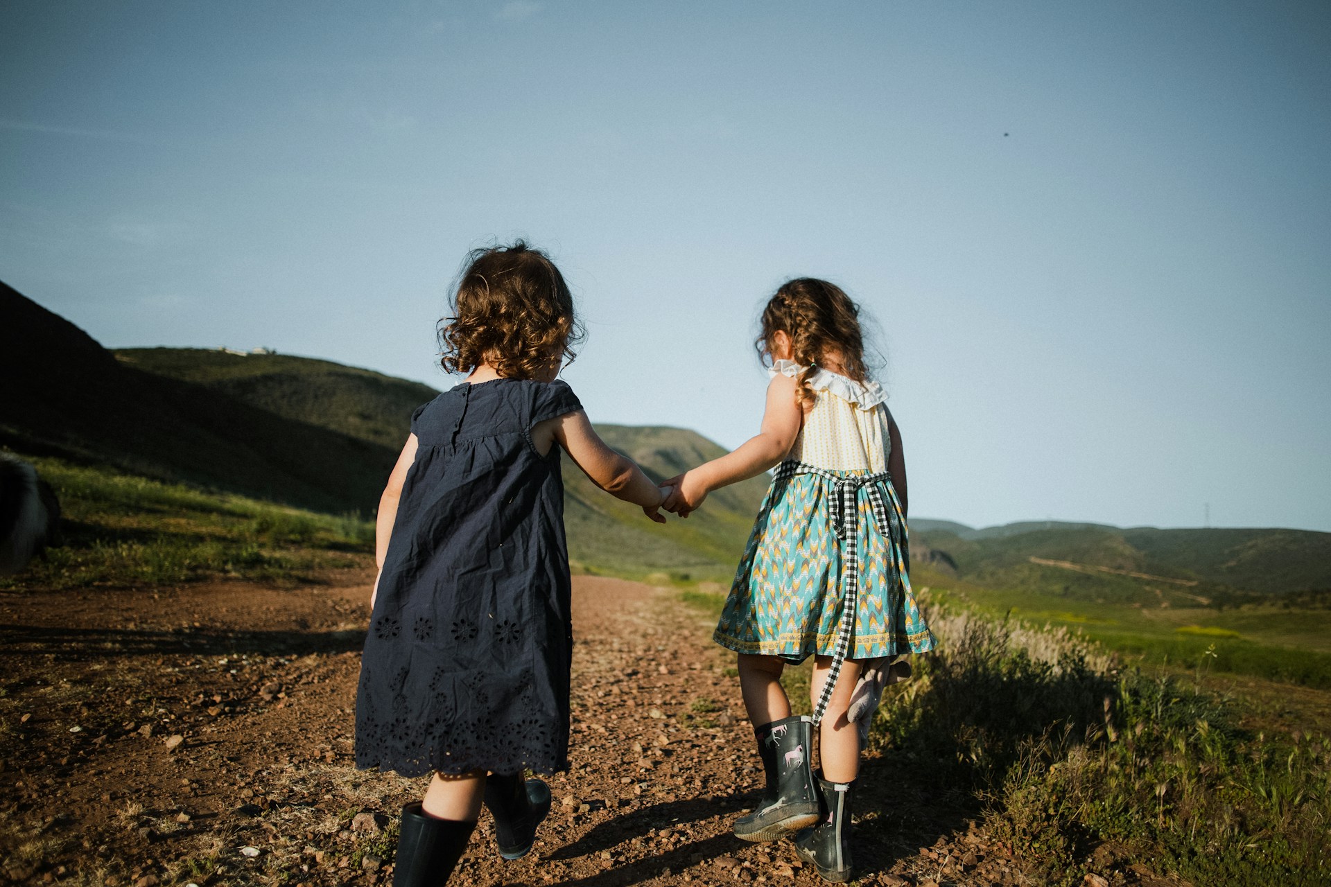 Two little girls from the back both dressed in sundresses and rubber boots are holding hands and walking along a dirt trail winding through grass-covered hills, representing dependant support claims.