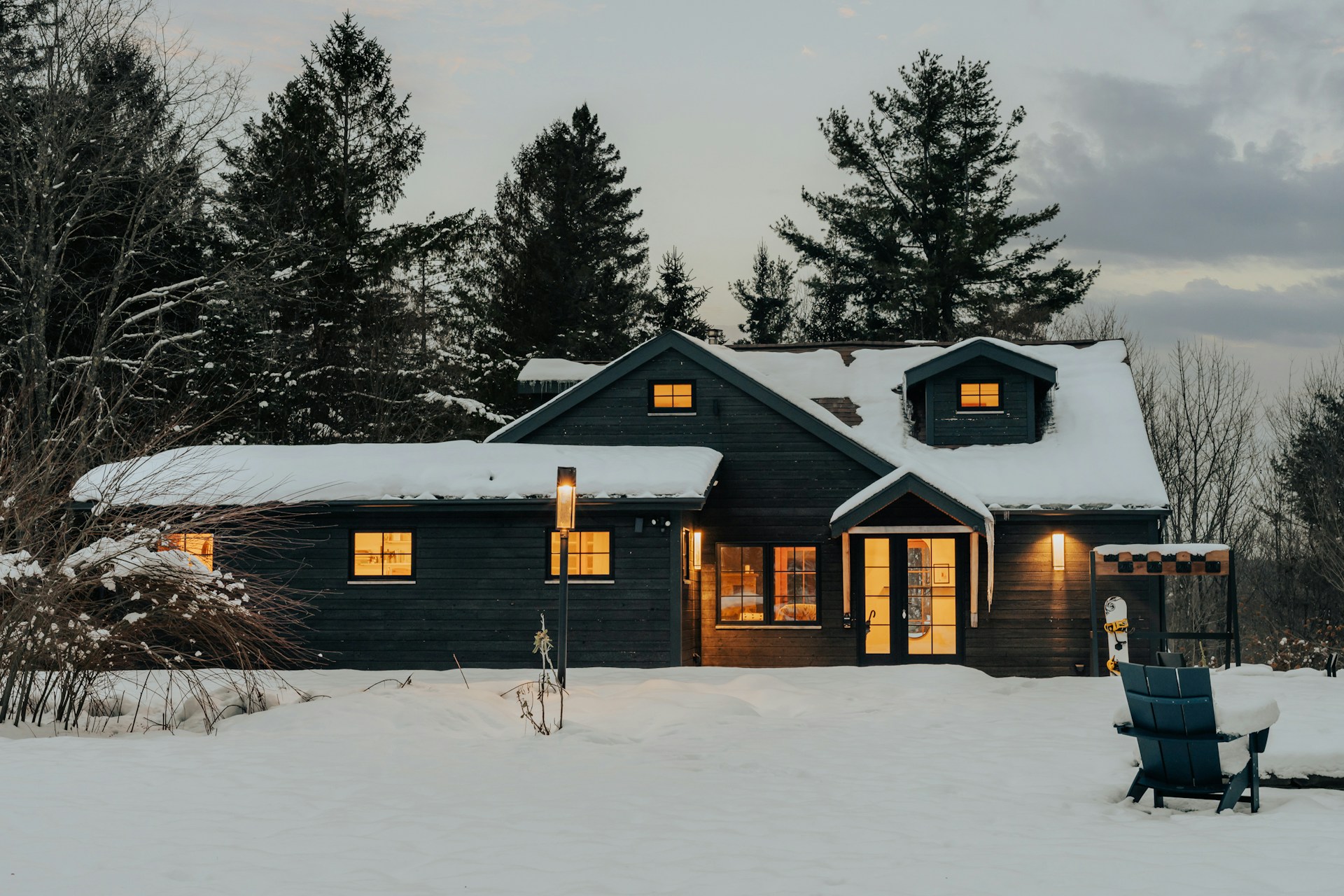 A large country house with dark wood siding, warm lighting spilling out from the windows, tall pine trees rising up behind it, and snow covering the lawn and roof, representing dependant support claims.