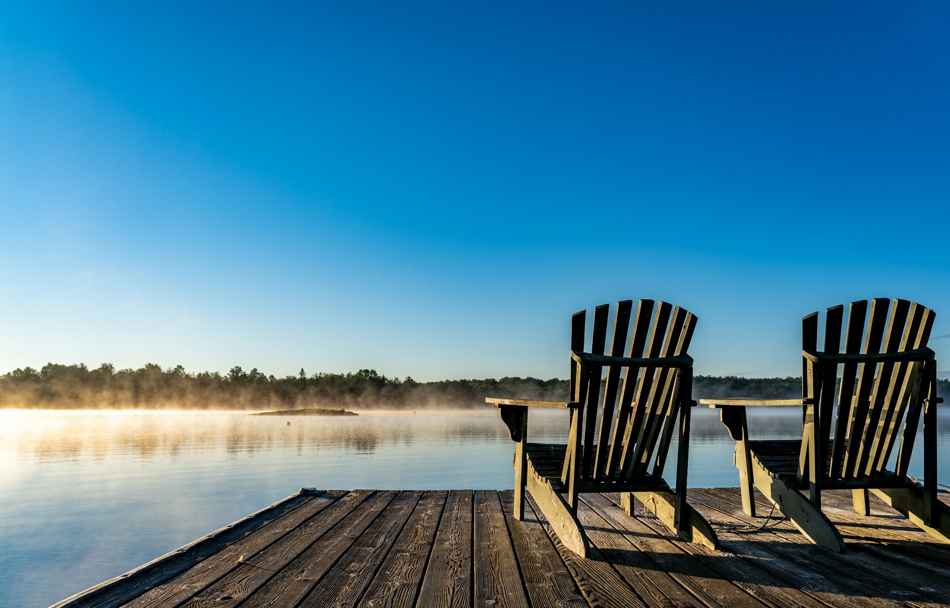 Two Muskoka chairs at the end of a dock look out over smooth water with mist drifting over the lake under a brilliant blue sky, representing family cottage trust disputes.