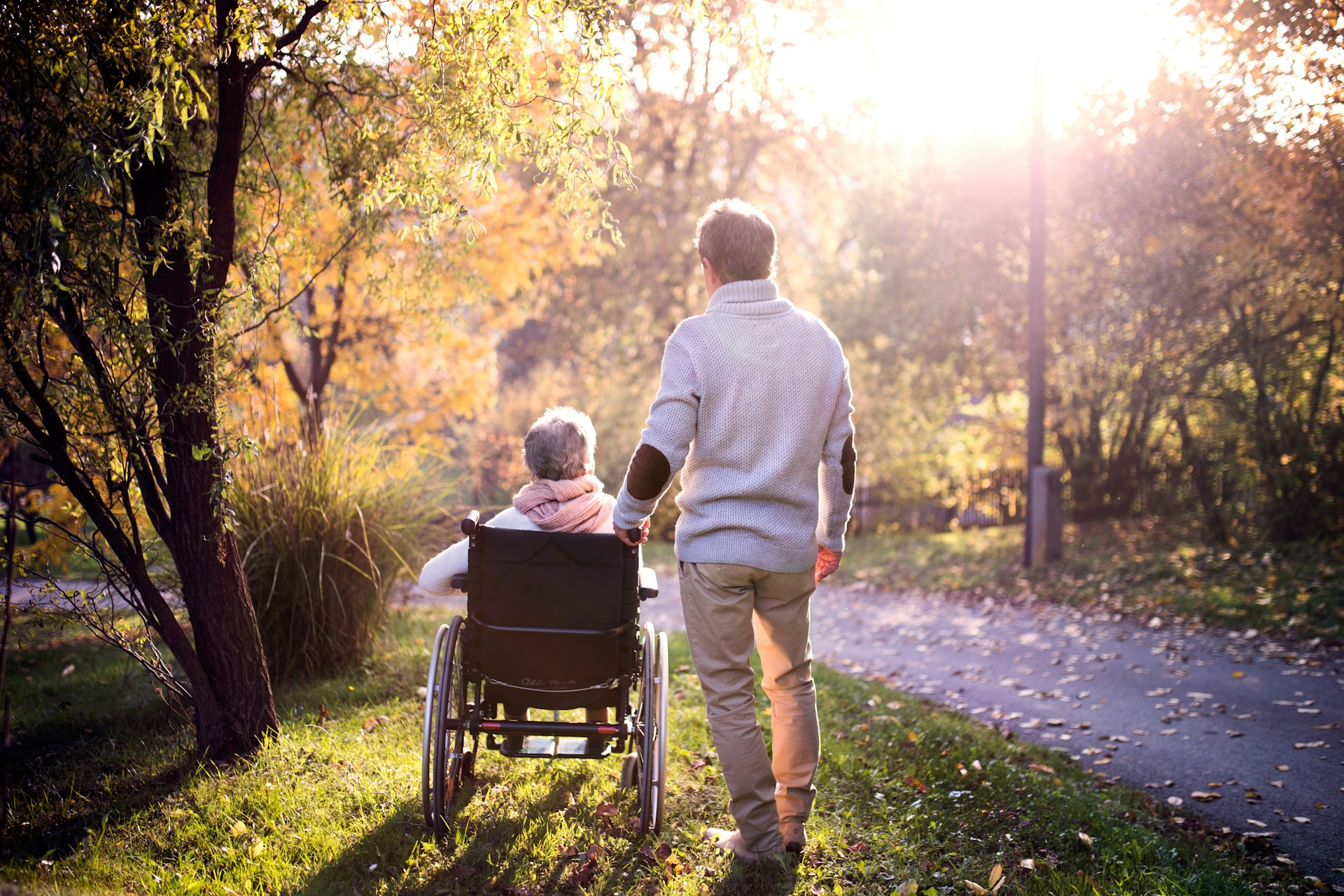 A man stands behind an elderly woman in a wheelchair next to a paved path dotted with autumn leaves while the sun casts a golden glow over the trees and bushes edging the path, representing capacity assessments and Ontario estate law.